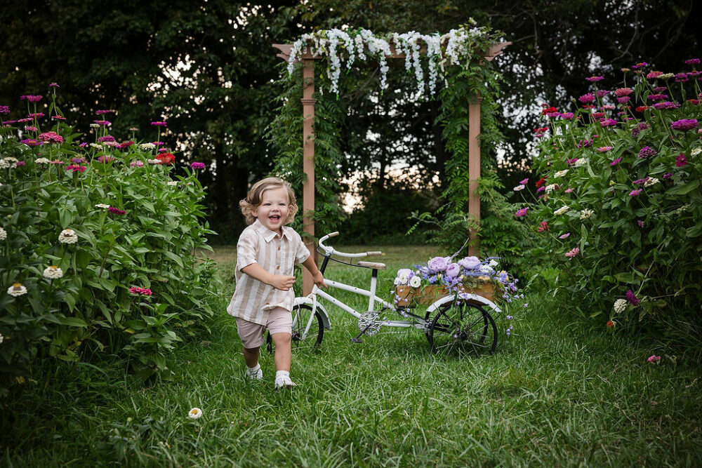 A cute photo of a boy running through a flower garden with a little bike in the background for flower garden mini sessions in Deptford, New Jersey.