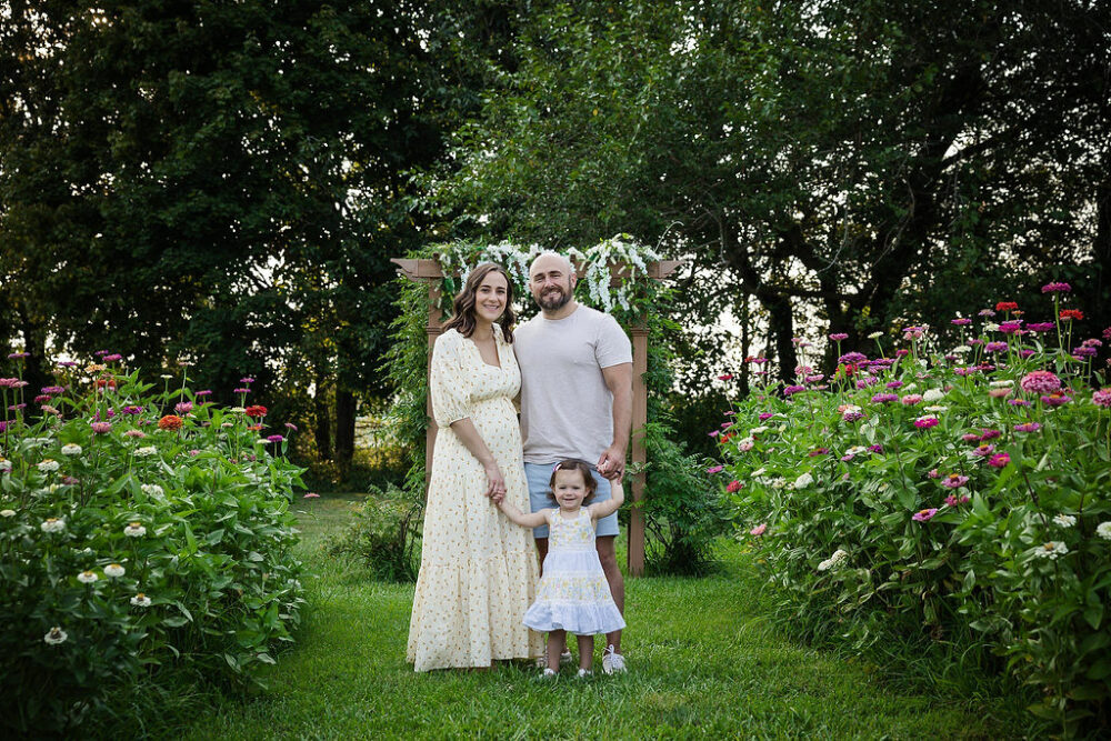 A family portrait of a woman and her daughter wearing a dress standing next to a man who is wearing a shirt and shorts, smiling for their outdoor family photoshoot in medford, New Jersey.