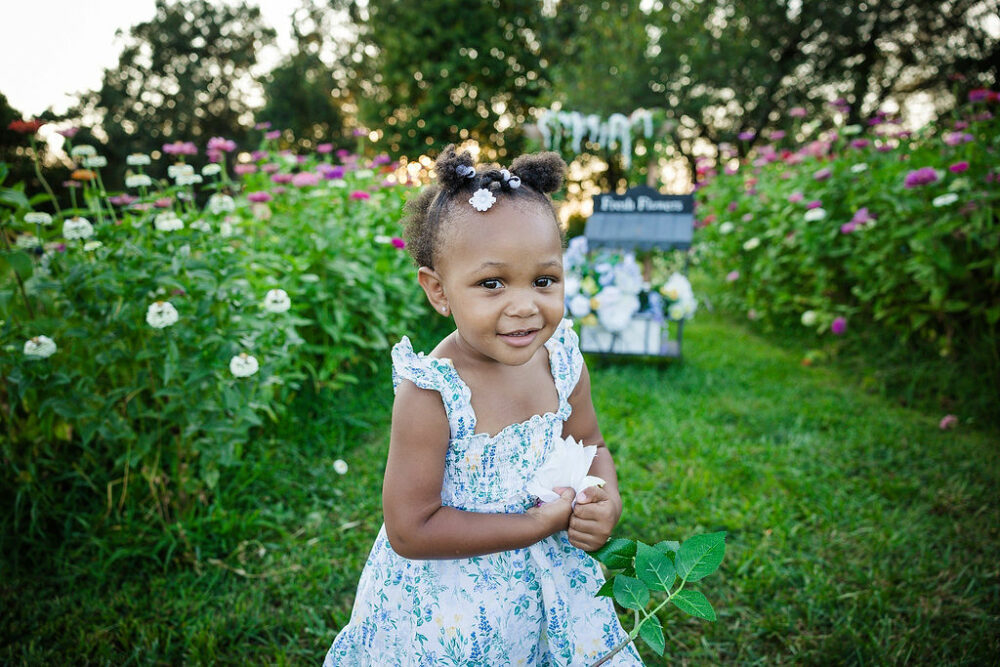 A close-up of a toddler girl wearing cute dress and hair ties, holding a flower as she stands and smiles for her flower garden mini sessions in Pemberton, New Jersey.