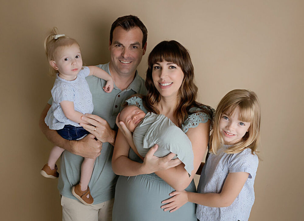 A family portrait of a man and woman standing next to each other posing with their three children against a neutral backdrop for their sons newborn photoshoot in Westampton, New Jersey.