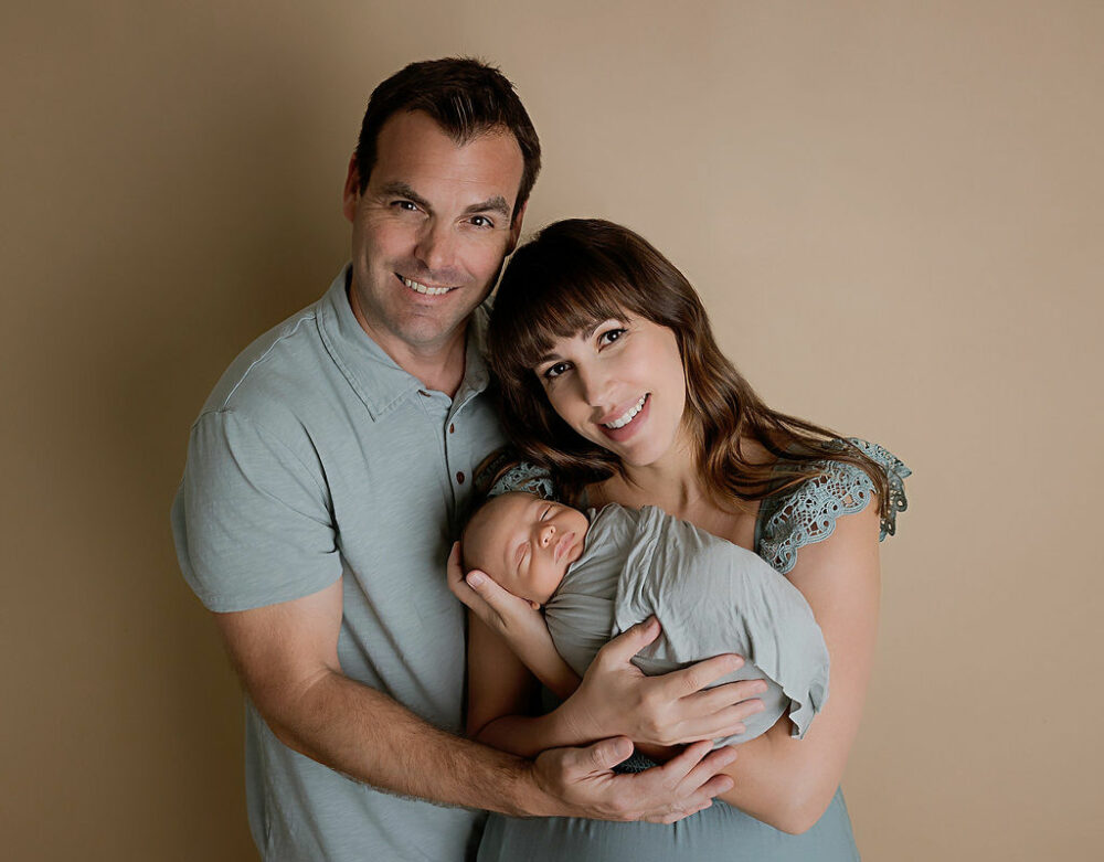 A portrait of a man and a woman standing next to each other hugging while she holds their a newborn son in her arms against a neutral backdrop for their sage green newborn session in Eastampton, new jersey.