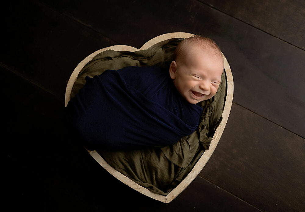Close-up of a newborn baby smiling in his sleep swaddled and placed in a heart shape photography prop for his professional newborn session in Medford, New Jersey.