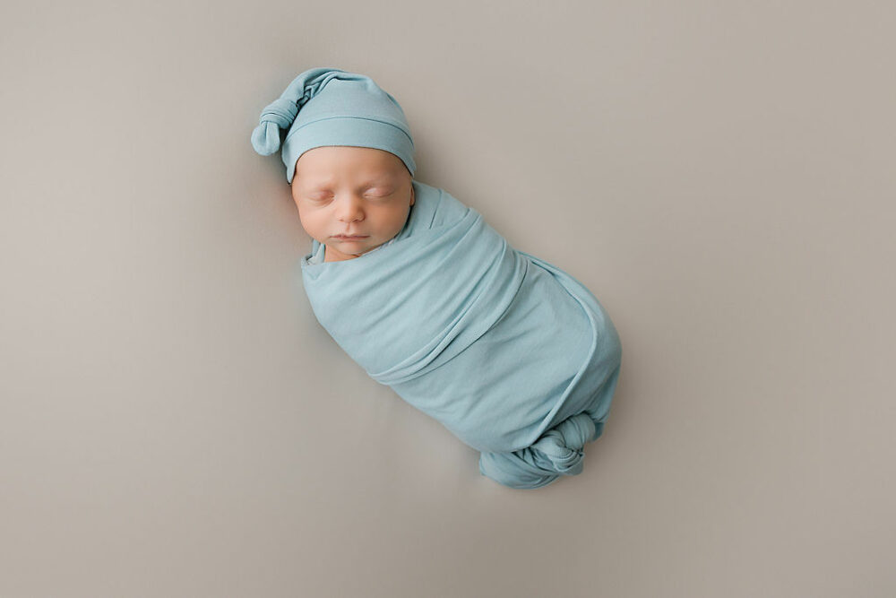 An infant portrait of a sleeping boy resting on his back and swaddled and wearing a cute hat for his sage green newborn session in Pemberton, New Jersey.