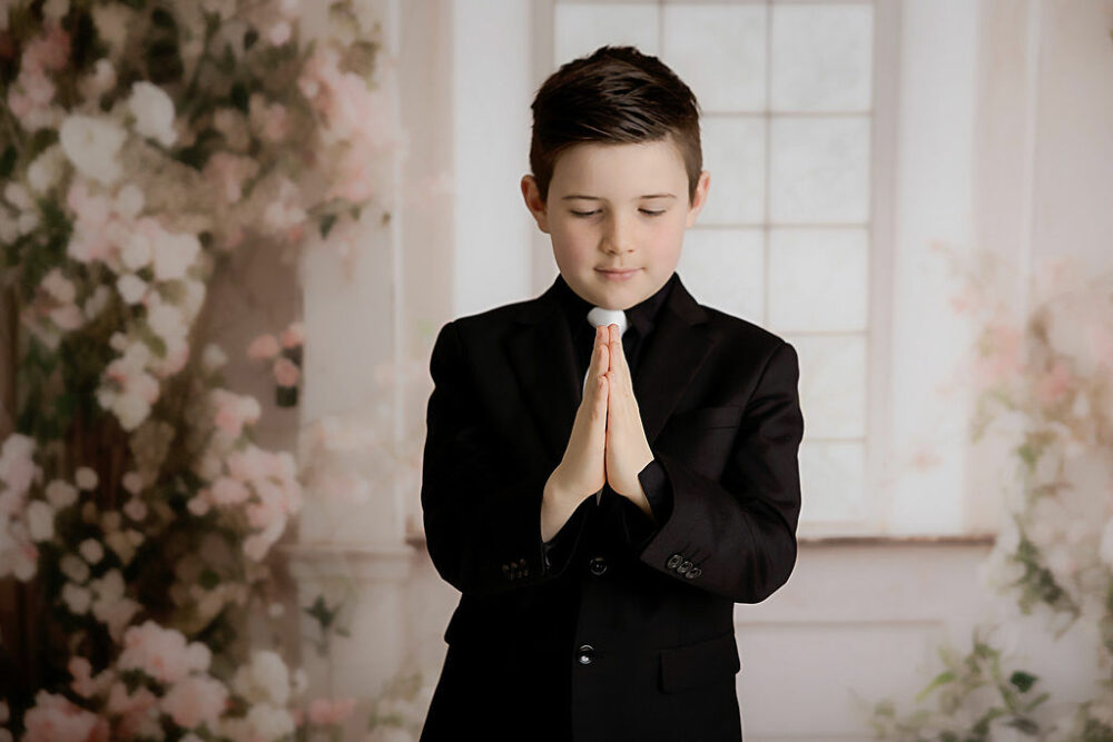 A portrait of a boy wearing black suit with white tie looking at his prayer hands against a white and pink backdrop for communion mini session in Delran, New Jersey.