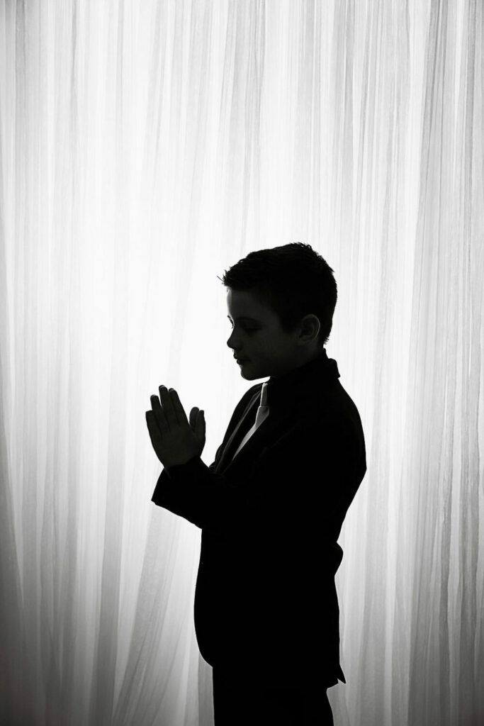 A black-and-white portrait of a boy standing sideways and holding his hands in prayer against a light backdrop for his communion mini session in Medford, New Jersey.