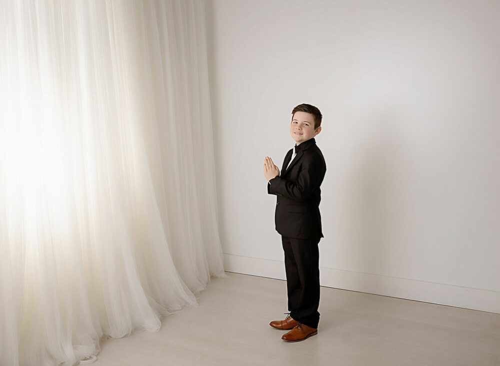 A photo of a boy smiling at the camera wearing a suit and holding prayer hands against a white background for his professional mini session in Moorestown, New Jersey.