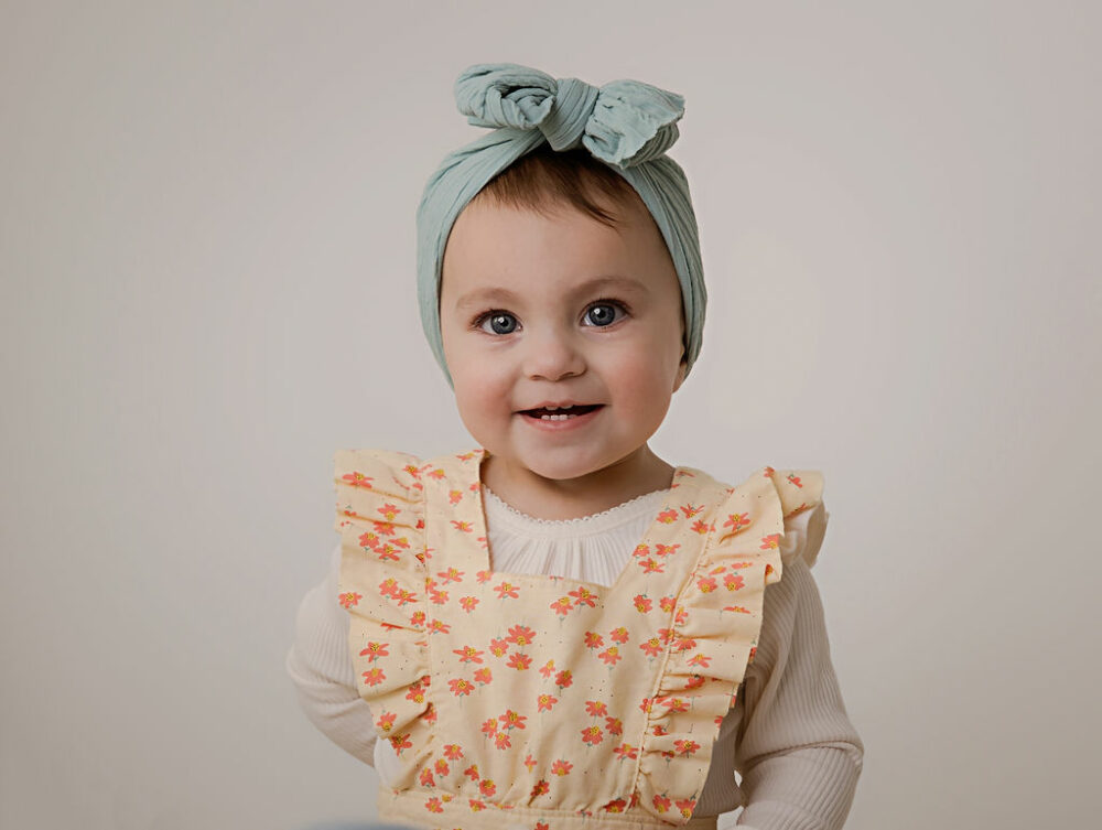 A close-up of a toddler girl smiling and wearing dress and cute headband during her simple first birthday session in Medford, New Jersey.