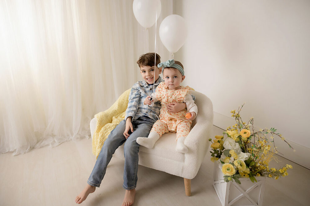A sibling portrait of a baby girl and her big brother sitting on a tiny couch against a bright backdrop in professional photography studio holding a balloon for her simple first birthday session in Deptford, New Jersey.