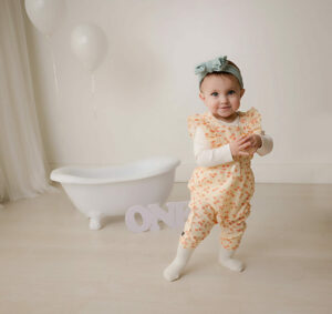 A cute photo of a toddler girl standing in front of tiny bathtub wearing cute overalls and headband, smiling for her simple first birthday session in Westampton, New Jersey.