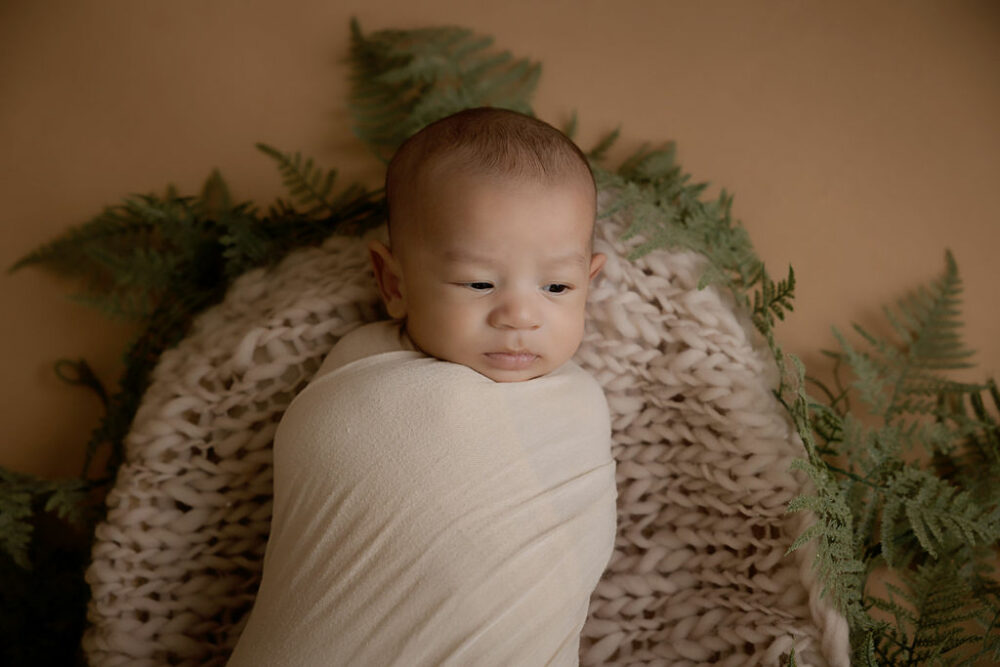 An infant photo of a boy in a swaddle, resting on blanket on top of photography prop for his classy newborn session in Deptford, New Jersey.