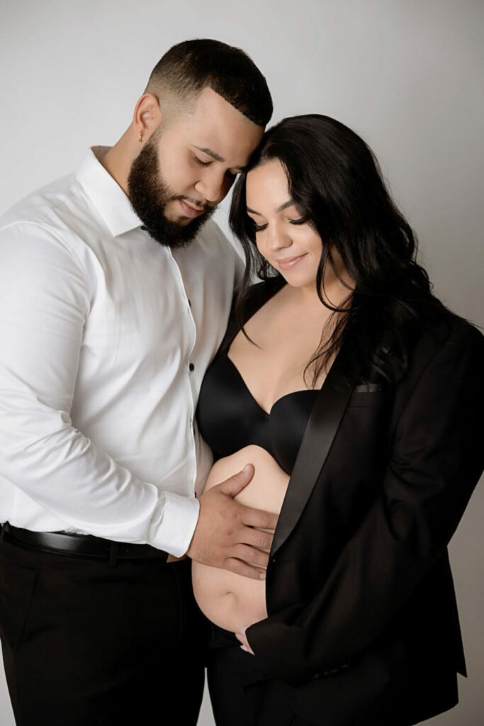 A beautiful photo of a man and a woman standing next to each other while they both touch her belly for their classy maternity session in Westampton, New Jersey.