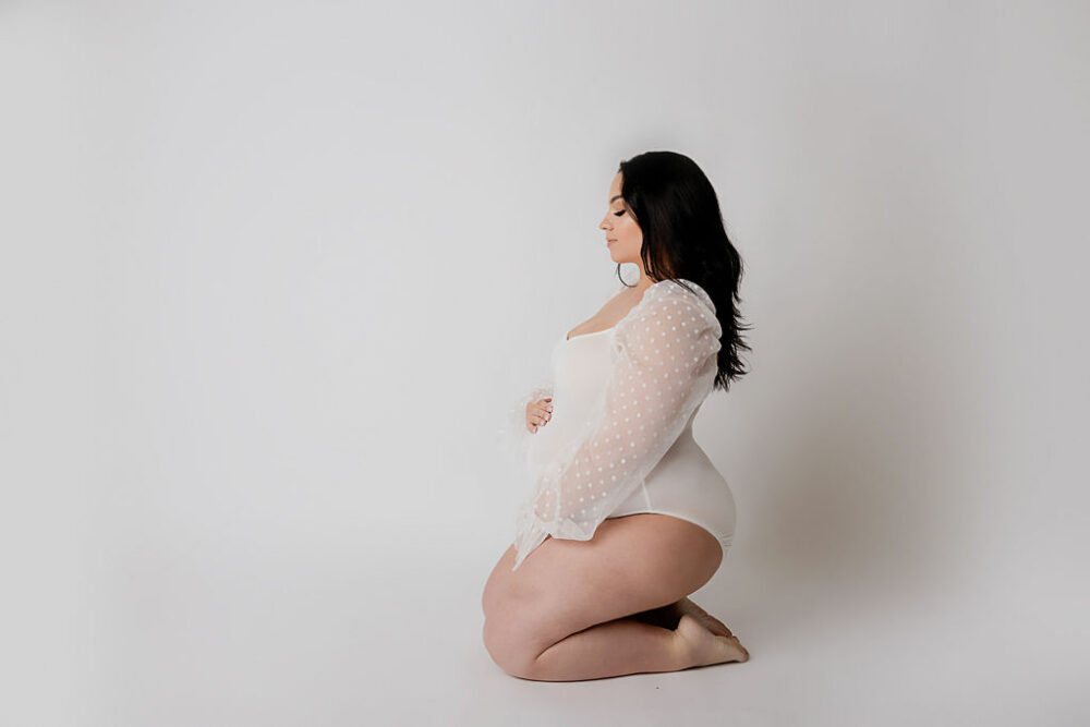 A beautiful photo of a woman kneeling and siting sideways and holding her belly, wearing a leotard with her hair down for her professional classy maternity session in Southampton, New Jersey.
