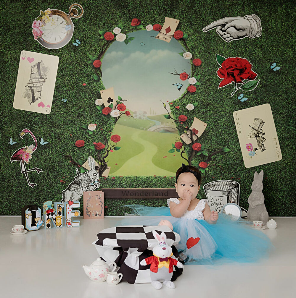A cute photo of a girl sitting on the floor next to cake stand against a Alice in wonderland backdrop with large keyhole adorned in greenery for her cake smash first birthday photoshoot in Eastampton, New Jersey.