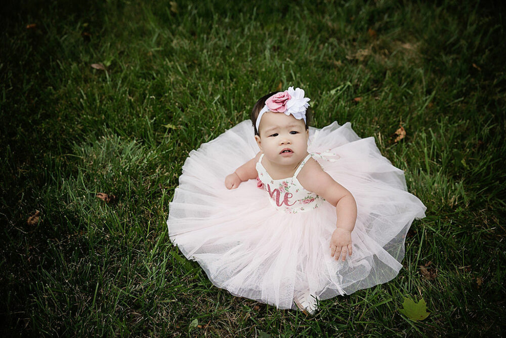 An infant portrait of a girl siting on grass and wearing a dress with the word one on it and a headband, looking up for her first birthday session in Southampton, New Jersey.