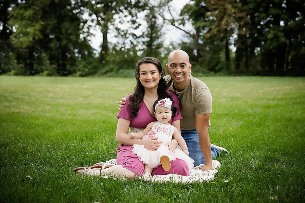 A beautiful family portrait of a couple and their daughter sitting outdoors, on grass, for their family photos in Camden, New Jersey.