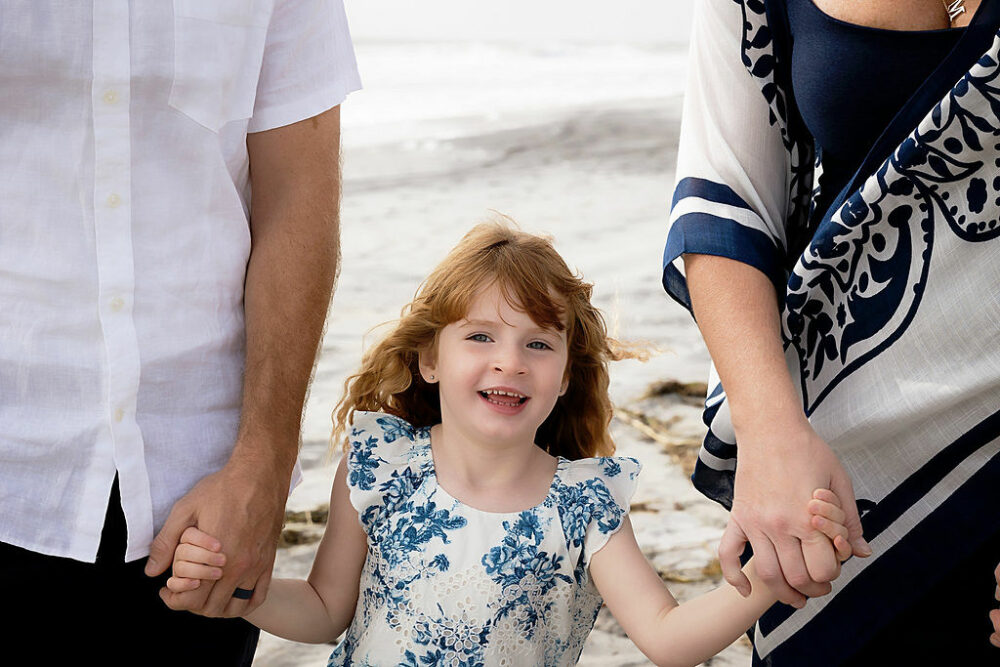 A close-up of a toddler girl smiling as she's holding her parents hands at the beach for their family photoshoot at the beach in Southampton, new Jersey.