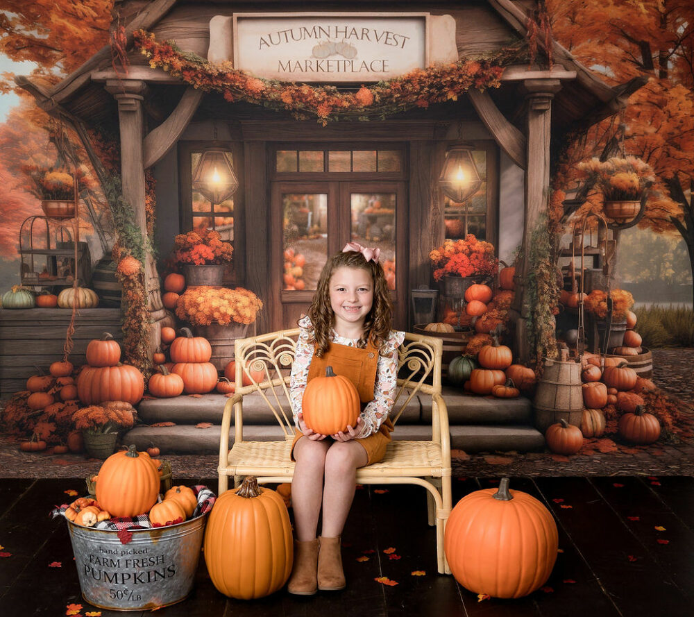A picture of a girl sitting tiny bench in front of a fall theme backdrop, holding a pumpkin and wearing overalls and boots for her fall in-studio mini session in Southampton, New Jersey.