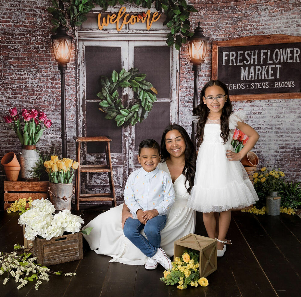 A cute family portrait of a woman and her two children posing against a market backdrop with flowers, smiling, and sitting for a Spring In-Studio Mini Session in Southampton, New Jersey.