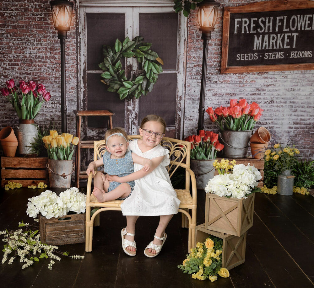 Cute picture of the two sisters sitting on tiny bench against a marketing backdrop with a ton of flowers for their spring in studio mini session in Westampton, New Jersey.