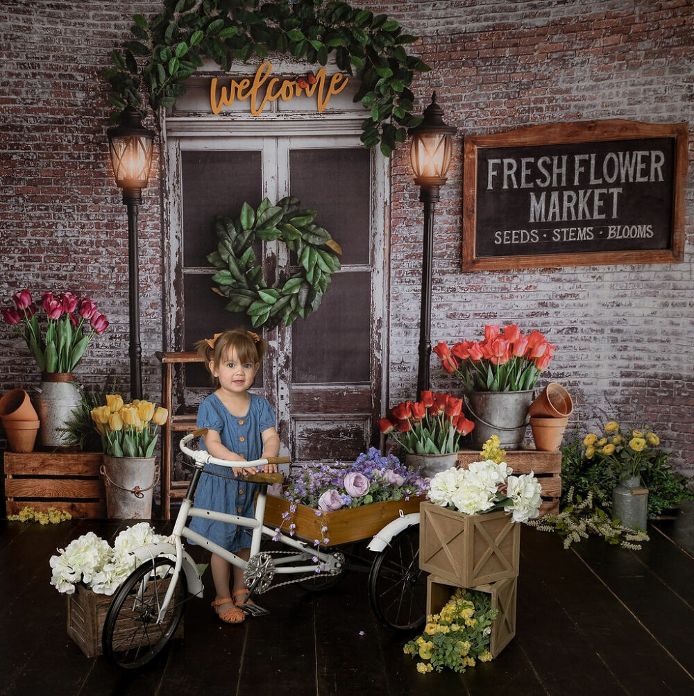 A toddler girl standing behind a bike photography prop adorned with flowers against a festive backdrop for her mile stone photography in Eastampton, New Jersey.