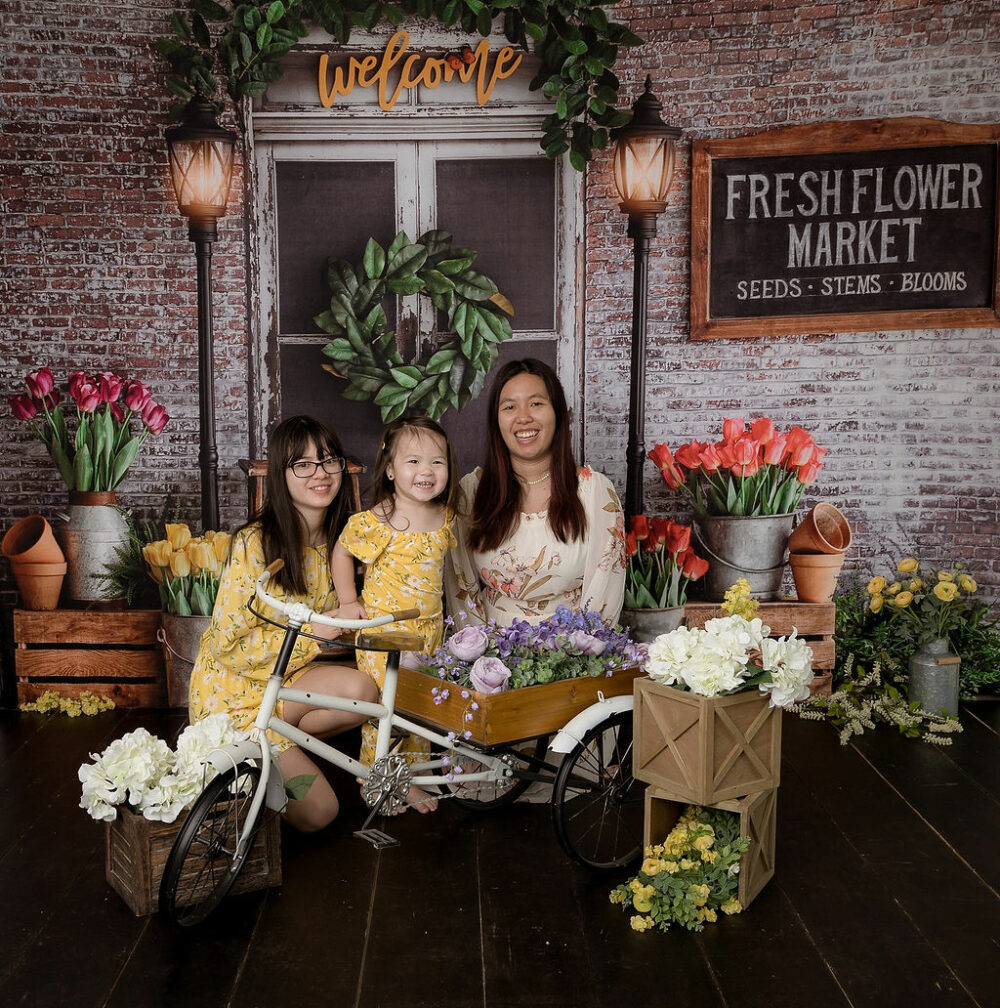 A family portrait of a woman and her two daughters closing behind a bike photography prop adorned with flowers for their spring in-studio mini session in Deptford, New Jersey.