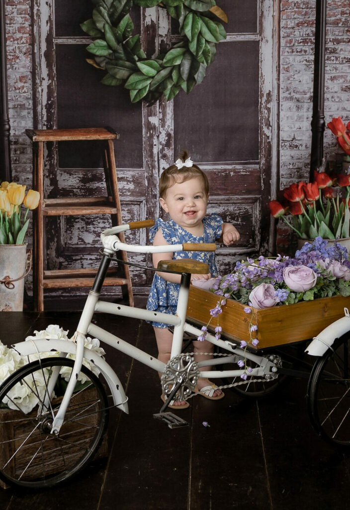 A baby portrait of a girl wearing a dress in a bowtie standing next to tricycle adorned with flowers against a seasonal backdrop for her monthly spring pics in Cherry Hill, new Jersey.