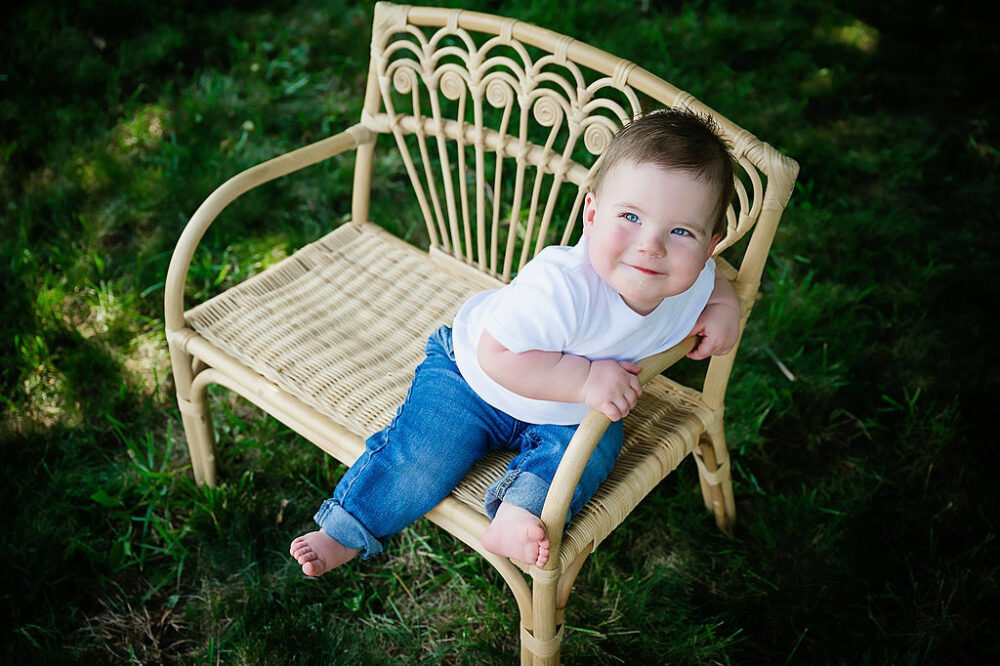 A portrait of a baby boy wearing jeans and t-shirt, sitting on tiny bench, outdoors, and smiling for their country first birthday pics in Southampton, New Jersey.
