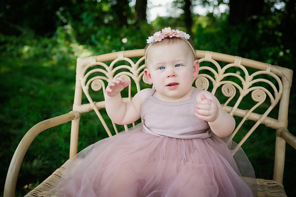 A one year old girl sitting on tiny bench wearing a cute dress and headband :-) for her outdoor baby milestones photo shoot in Westampton, New Jersey.