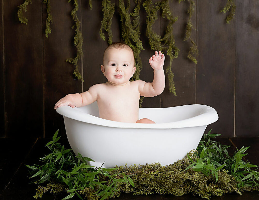 A baby in a tiny bathtub photography prop waving while sitting, adorned with greenery for his fishing first birthday session in Southampton, New Jersey.