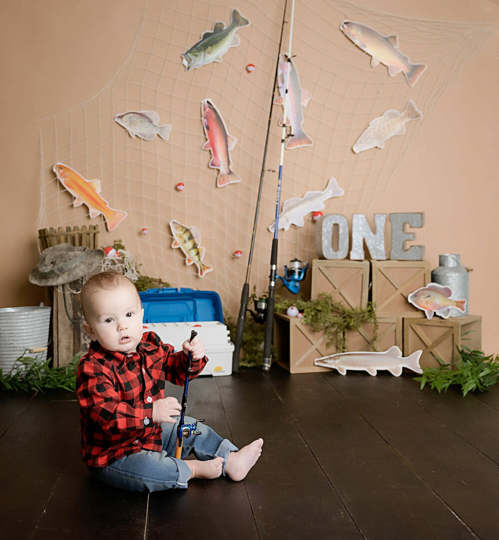 Baby photography of a toddler boy wearing jeans and button up shirt, sitting not he ground holding. sighing pole for his Fishing first birthday session in Eastampton, New Jersey.
