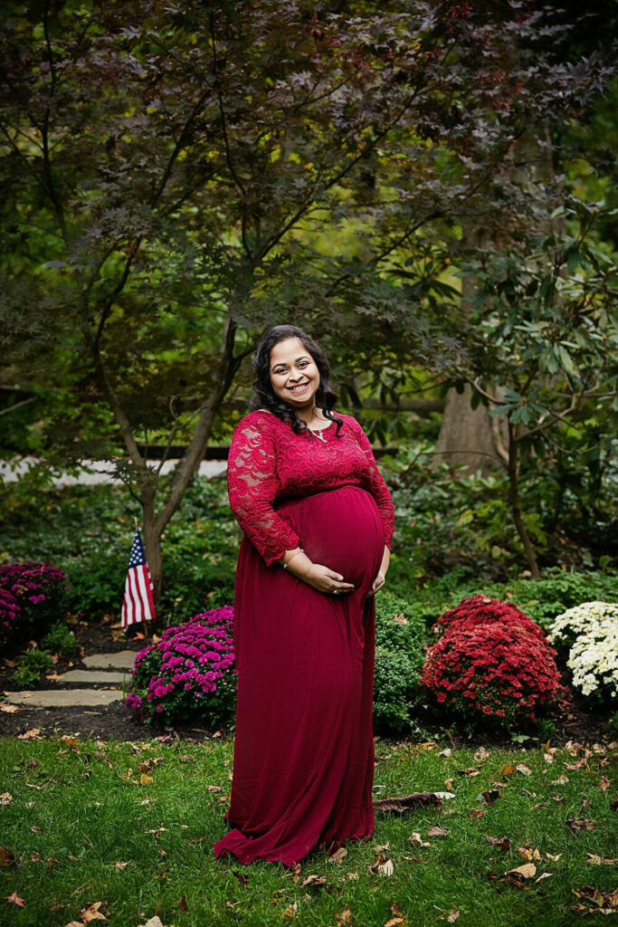 A woman standing outdoors against rosebushes wearing maternity gown and holding her belly while smiling for her maternity photo session in medford, New Jersey.