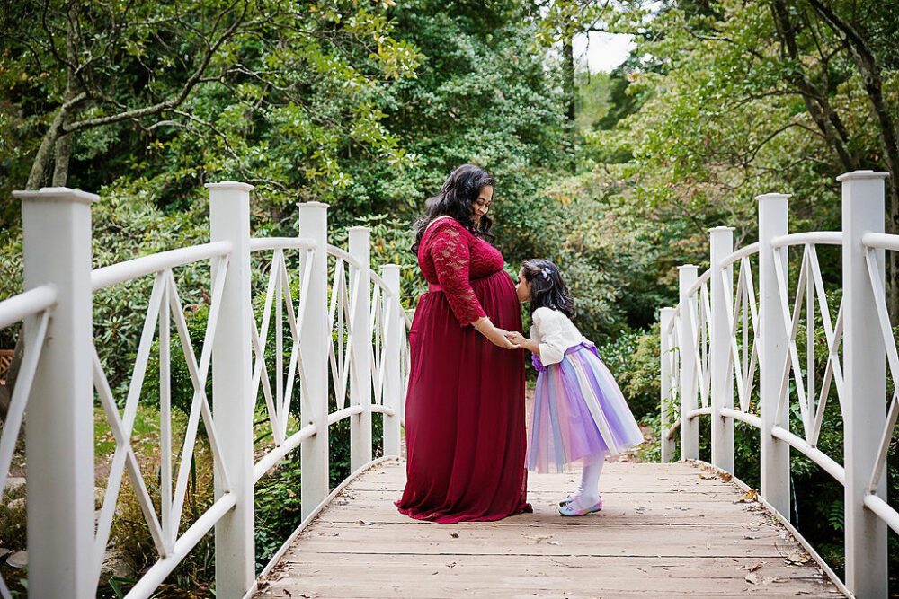 A cute mommy portrait of a woman holding hands with her toddler daughter as she kisses her belly, standing on a bridge against lush greenery for her maternity session in Hamilton, New Jersey.