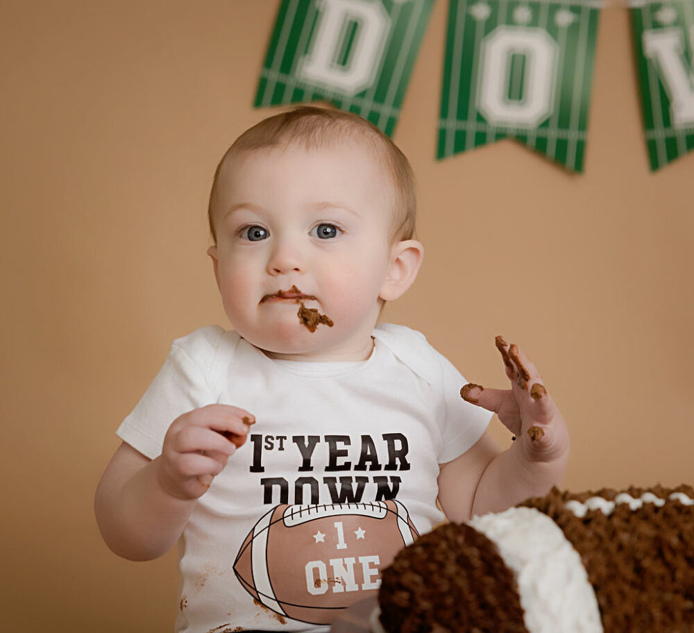 A cute photo of a boy eating cake during his cake smash photography near me with a custom first down first birthday session backdrop in professional studio in Medford, New Jersey.