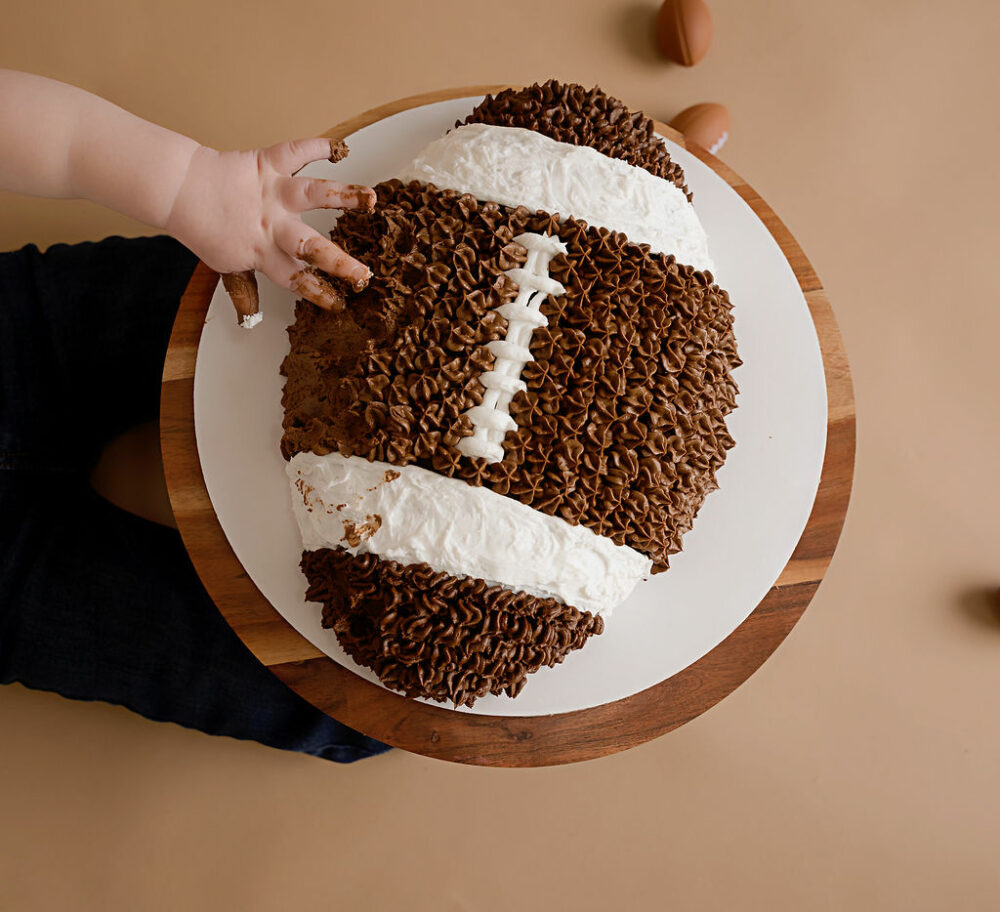 A photo of a toddler hand grabbing icing off of his football-shaped cake during his first down first birthday session in Pemberton, New Jersey.