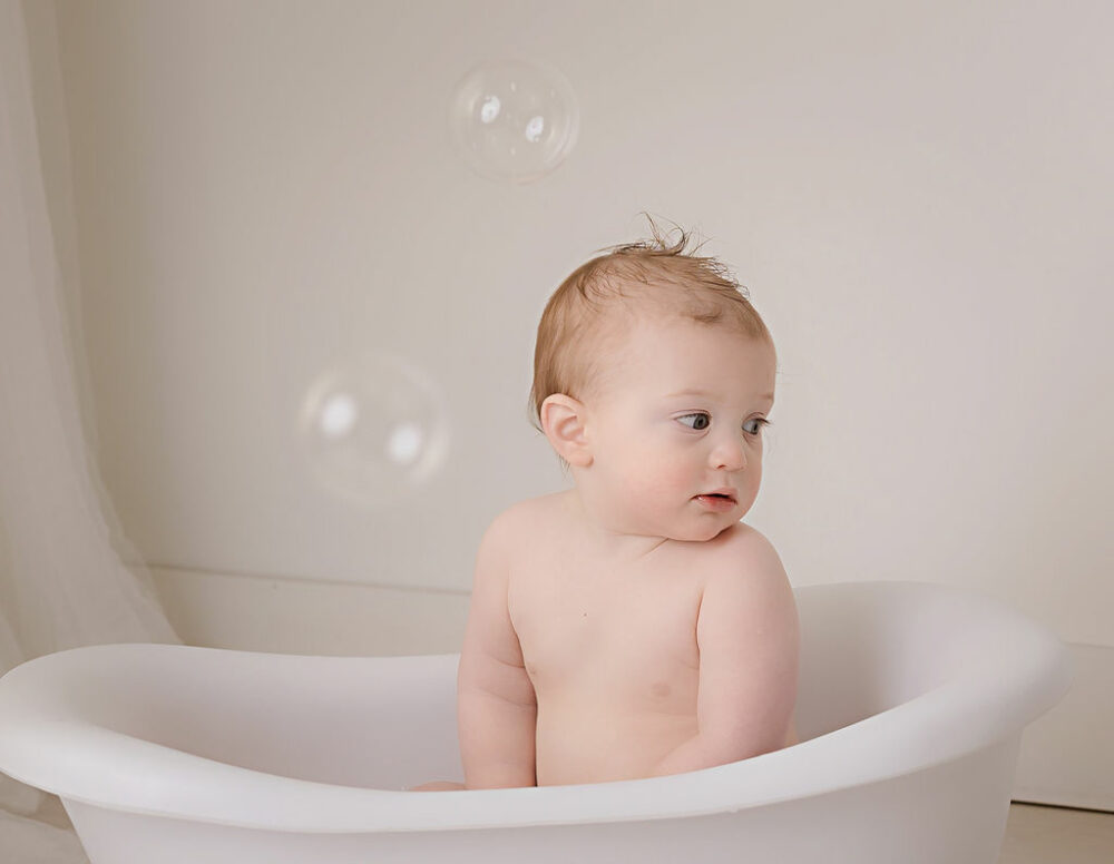 A cute photo of a boy sitting in a little bathtub photography prop looking at bubbles during his first down first birthday session in Eastampton, New Jersey.
