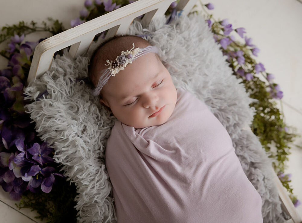 A close-up of a sleeping baby girl placed on tiny crib photography prop lined with fuzzy blanket and purple flowers for her lavender newborn session in Medford, New Jersey.
