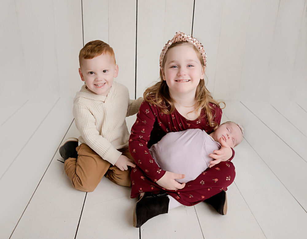 A cute picture of three siblings sitting on the floor in a professional studio smiling for their little sisters newborn baby pics taken and professional studio for her lavender newborn session in Westampton, New Jersey.