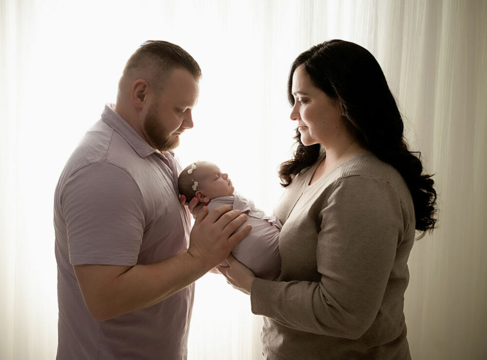 A family portrait of a man and woman holding their baby between them as they look down at her while she sleeps in their arms against a light and bright backdrop for their child's lavender newborn session in Southampton, New Jersey.