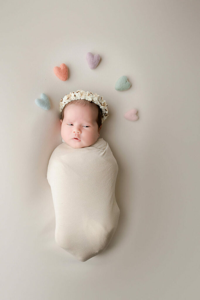 An infant portrait of a girl swaddle and wearing flower headband adorned with felt harts post for her professional newborn photography session in Medford, New Jersey.