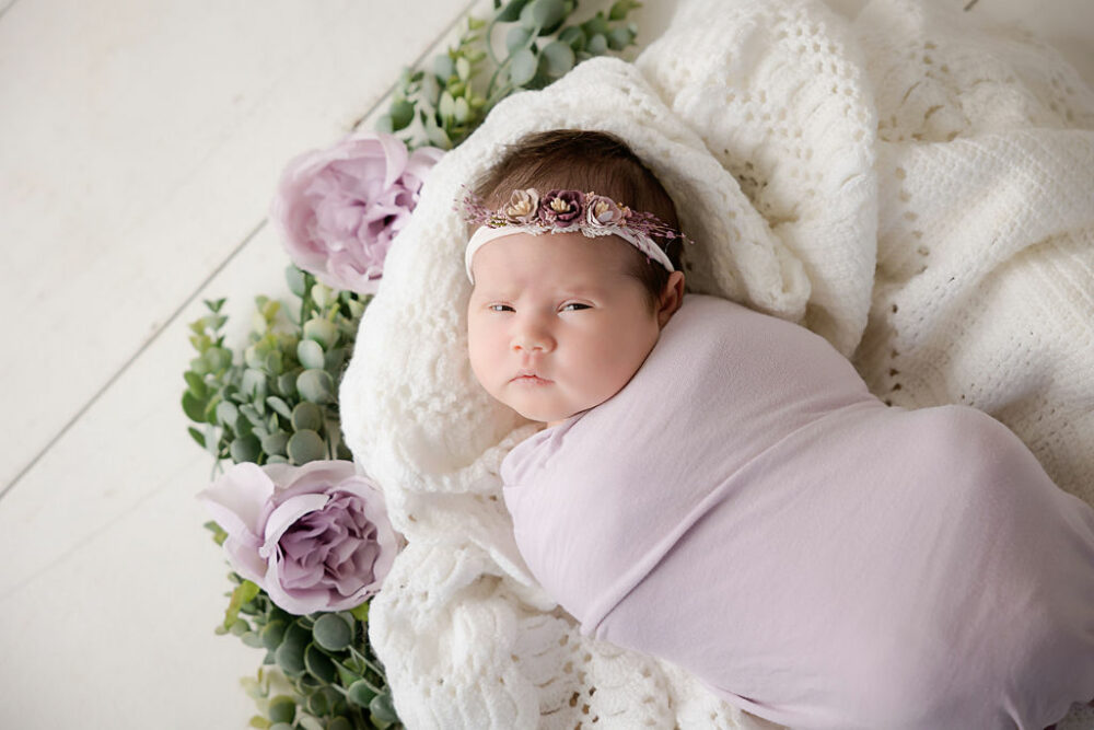 Take a picture of a newborn girl resting on her back on blanket adorned with greenery and flowers and wearing a headband for her professional newborn session in pemberton, New Jersey.