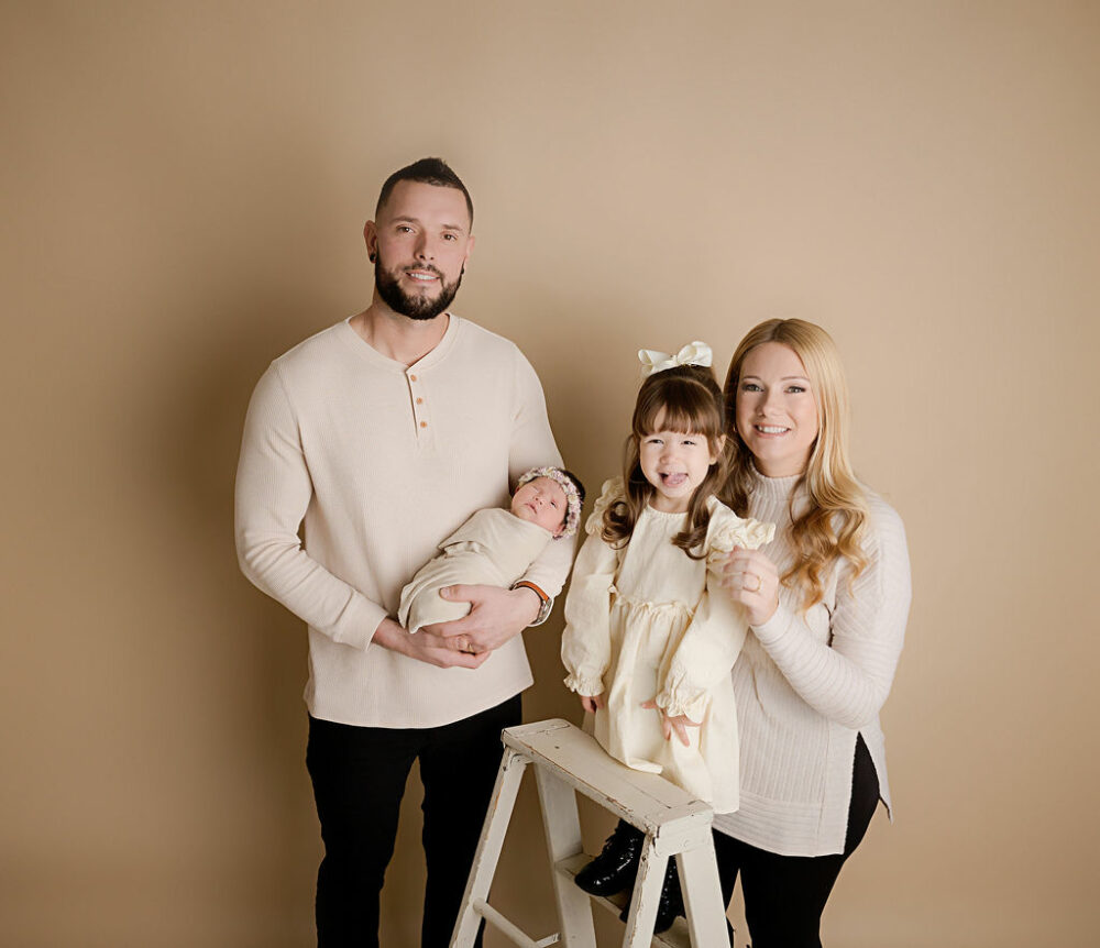 A beautiful family portrait of a man and woman posing and holding their infant daughter and toddler daughter wearing matching clothing for a formal newborn session in Tabernacle, New Jersey.