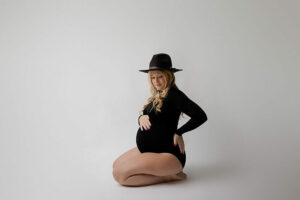 A woman sitting on the floor with her legs tucked in posing for her formal maternity photo shoot wearing one piece and hat for her pregnancy portraits in Eastampton, New Jersey.