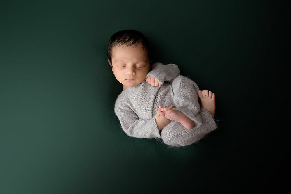 A portrait of a boy sleeping on his back wearing a one piece with his legs tucked in on a solid backdrop for his cute newborn session in Southampton, New Jersey.
