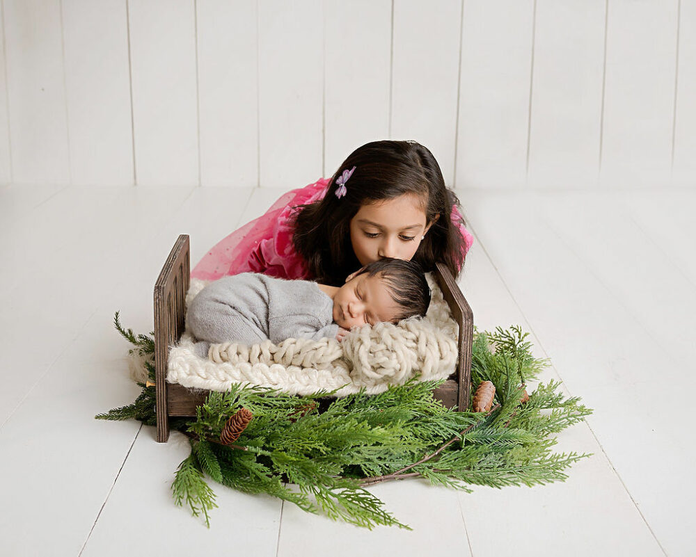 A sibling portrait of a big sister kissing her little brother as he sleeps on tiny photography prop for bed during his newborn portrait studio in Westampton, New Jersey.