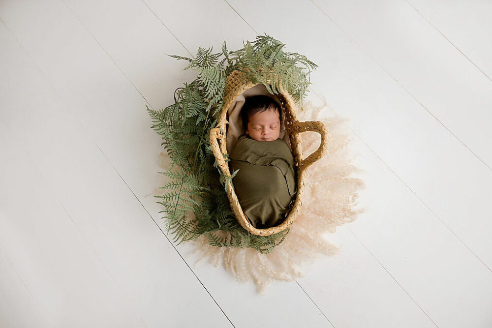 A baby portrait of a newborn wrapped and placed carefully in a woven tote adorned with greenery for his professional baby pictures in Eastampton, New Jersey.