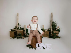 An adorable photo of a boy sitting on a tree stump photography prop with greenery and trees, smiling for his happy camper first birthday session in Delran, New Jersey.