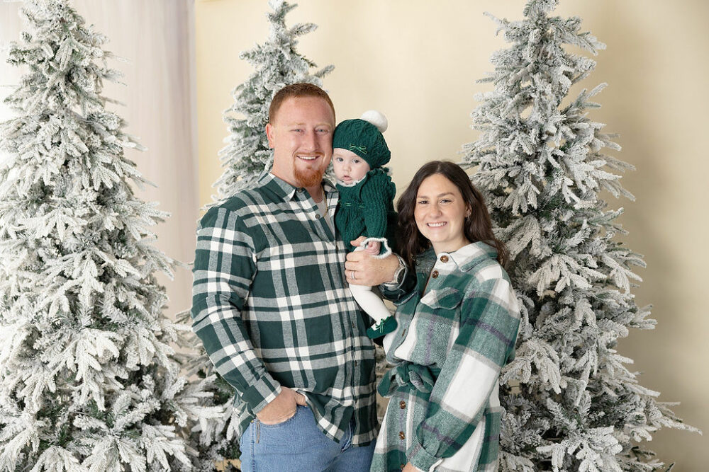 A beautiful photo of a man and woman standing side by side and holding their daughter between them for their green holiday mini sessions in Westampton, New Jersey.