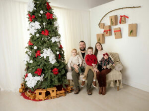 A beautiful formal portrait of a man and woman sitting with their three children on a sofa next to large Christmas tree fro their holiday mini Session in Eastampton, New Jersey.