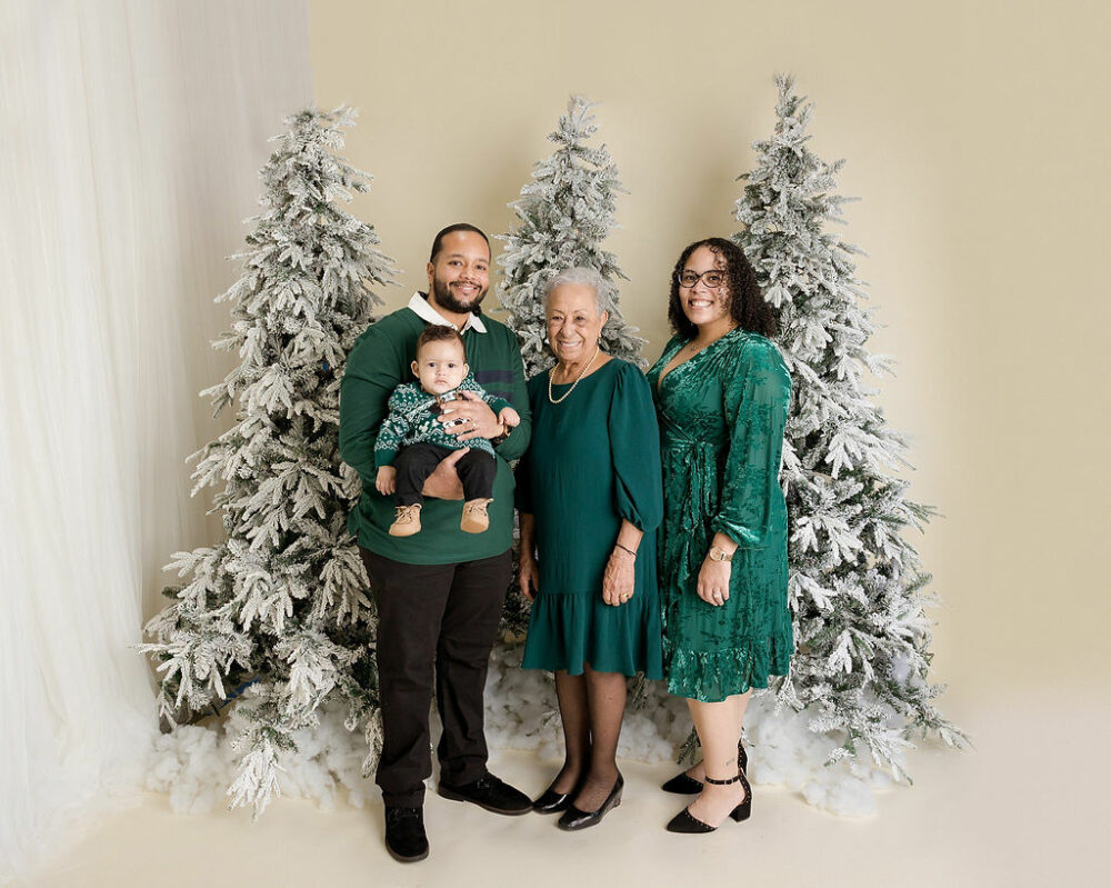 A beautiful portrait of a man and woman posing with their son and his grandmother in front of frosted trees for their holiday mini session in professional studio in Medford, New Jersey.