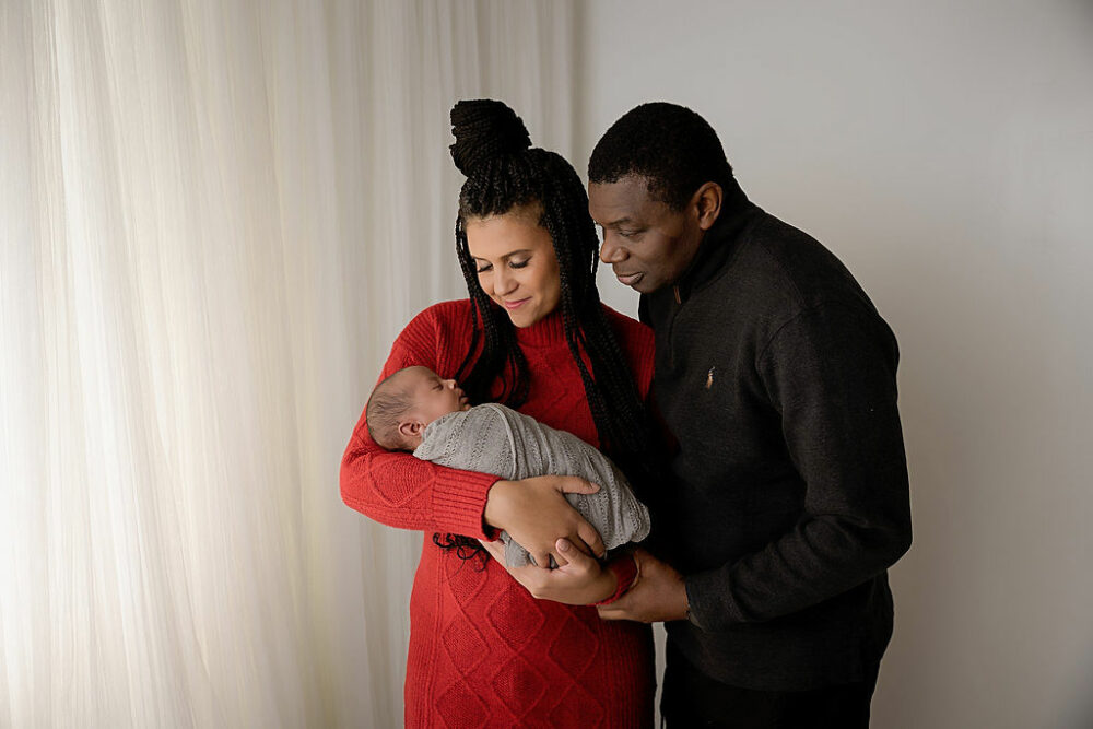 A man and woman standing next to each other and looking at their baby against the light and bright backdrop wearing moody colors as they hold their son for a red newborn session in Eastampton, New Jersey.